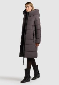 Dugi, podstavljeni kaput u tamno sivoj boji, sa visokim kapuljačom, prednjim patent-zatvaračem i bočnim džepovima. Uparen s crnim čizmama na potpeticu do gležnja.