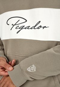 Καφέ και λευκή φούτερ μπλούζα με το κείμενο "Pegador" σε μαύρη καλλιγραφία, και ένα λογότυπο κεντημένο στο μανίκι. Μαλακό ύφασμα και ραμμένα μανίκια.