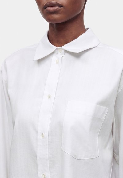 Gros plan d'une personne portant une chemise blanche boutonnée avec col et poche poitrine, montrée des épaules aux lèvres.