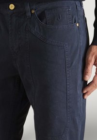 Primo piano di pantaloni blu navy con tessuto testurizzato, cuciture dettagliate, tasca frontale, bottone di ottone e una mano appoggiata vicino alla coscia.