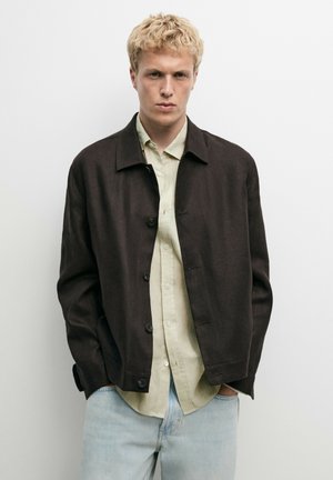 Junger Mann mit kurzen blonden Haaren, der eine dunkelbraune, geknöpfte Jacke, ein hellbeiges Hemd und hellblaue Jeans vor einem schlichten weißen Hintergrund trägt.