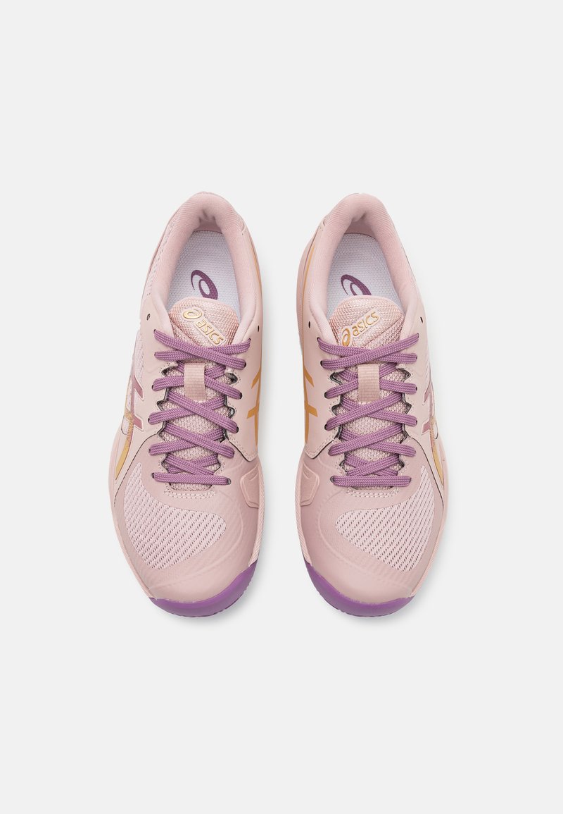 Zapatillas deportivas de color rosa claro con detalles y cordones morados. Textura de malla en los laterales, suela de goma y marca dorada en la lengüeta.