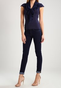 Marinblå blus med polka prickar och korta ärmar, prydd med en rosett, som matchas med mörkblå smala jeans. Modellen bär hudfärgade klacksandaler.