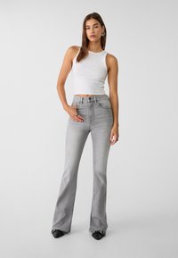 Jean grise évasée à taille haute, en denim. Associée à un débardeur blanc ajusté. Les accessoires incluent des boucles d'oreilles créoles et des chaussures noires.