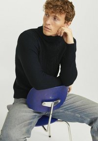 Pull col roulé en tricot torsadé bleu marine avec un motif texturé, porté avec un jean gris clair, assis sur une chaise bleue avec des accents en métal.