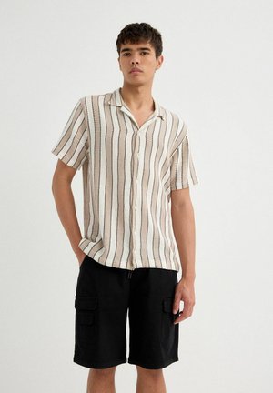 Camisa de botones de manga corta con rayas verticales en beige y azul marino, confeccionada en tejido texturizado; combinada con pantalones cortos cargo negros con bolsillos.