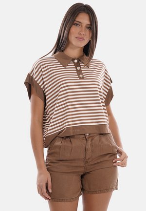 Donna che indossa una polo a maniche corte a righe marroni e bianche con bottoni e pantaloncini marroni a vita alta, in piedi davanti a uno sfondo bianco.