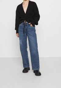 Svart stickad tröja med djup V-ringning och dragsko i midjan, tillsammans med löst sittande blå denimjeans och svarta kraftiga ankelstövlar.