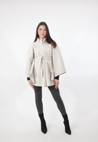 Kremowy płaszcz peleryna z wiązaniem w talii, wysokim kołnierzem i szerokimi rękawami, zestawiony z ciemnoszarymi dżinsami i czarnymi botkami na obcasie. Gładka faktura.