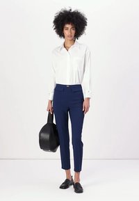 Λευκό πουκάμισο με κουμπιά και τσέπη, ναυτικές κομμένες παντελόνες και μαύρες λόφερ. Κρατώντας μια μαύρη τσάντα, στέκεται απέναντι από ένα ουδέτερο φόντο.