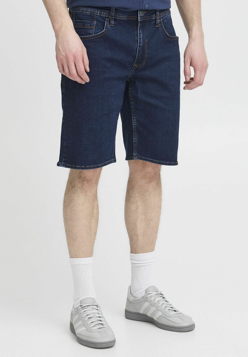 Shorts en denim bleu foncé avec une coupe droite, coutures visibles et cinq poches. Associés à des baskets grises et des chaussettes blanches.