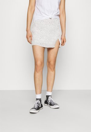 Mini-jupe blanche avec de petites taches noires, dotée d'une fente sur le côté, associée à des baskets montantes noires et des chaussettes blanches mi-mollet.