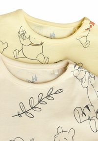 Deux t-shirts en coton jaune avec des illustrations de personnages de dessins animés, y compris Winnie l'Ourson et Tigrou, ainsi que des motifs de feuilles le long de l'encolure.
