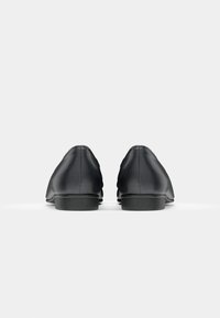 Chaussures en cuir noir avec une texture lisse, bout arrondi et semelles plates noires, vues de l'arrière. Design minimaliste, sans matériel visible.