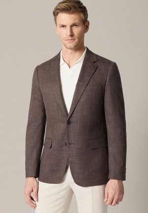 Mężczyzna w brązowej teksturowanej marynarce, białej koszuli i jasnych spodniach na jednolitym tle.