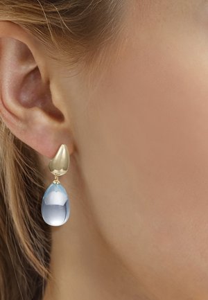 Goldener Ohrstecker in unregelmäßiger Form mit facettiertem, transluzentem, blauem Tropfenanhänger, getragen am linken Ohr einer Frau.