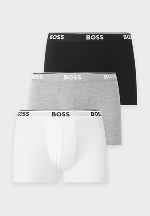 Uomo atletico dalla pelle scura in piedi a piedi nudi indossa boxer neri firmati BOSS su uno sfondo chiaro e uniforme.