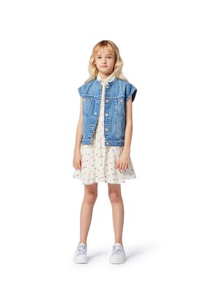 Veste sans manches en denim sur une robe blanche à motifs rouges ; modèle portant des baskets blanches, debout sur un fond uni.