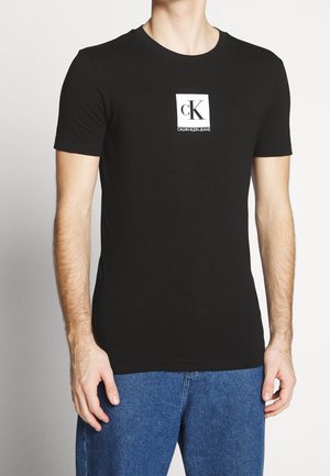 Svart t-shirt med korta ärmar av bomull, med en vit rektangulär logotyp där "cK" och "CALVIN KLEIN JEANS" är tryckta i svart.