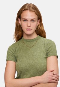 Groen top met korte mouwen en een gestructureerde brei, gecombineerd met een gouden ketting. Het model heeft golvend haar en draagt ringoorbellen.
