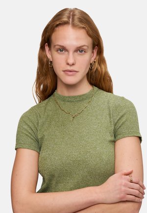 Grön kortärmad topp med en strukturerad stickning, ihop med ett guldhalsband. Modellen har vågigt hår och bär hängande örhängen.