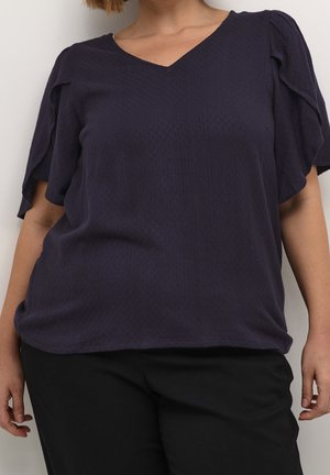 Femme portant un haut à manches courtes texturé violet foncé avec un col en V et un pantalon noir, sur un fond clair uni.