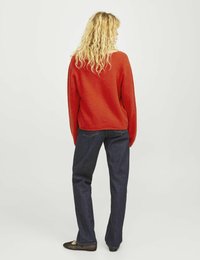 Roter Pullover mit lockerem Schnitt und strukturiertem Finish, kombiniert mit dunkelblauen, geraden Jeans und schwarzen flachen Schuhen.