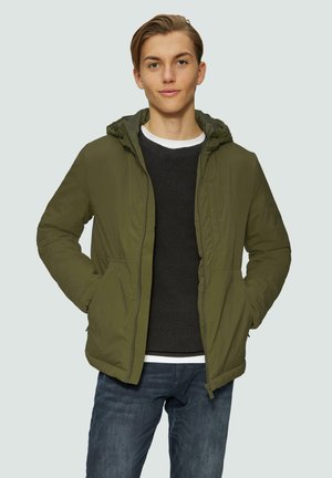 Veste à capuche vert olive avec un extérieur lisse, poches avant et fermeture éclair. Portée par-dessus un pull noir texturé et un jean bleu.