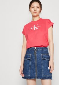 Raudonas medvilninis marškinėliai su suktomis rankovėmis ir "Calvin Klein Jeans" logotipu, derinami su mėlynu džinsiniu sijonu, turinčiu priekines užtrauktuko kišenes ir dvi kišenes.