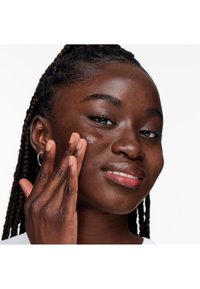 Ansiktspleie med en person som påfører en krem. Har fletter, glatt hud, og et subtilt smil. Lett sminke med varme toner.