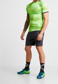 Camisa atletica de manga corta verde con diseño texturizado, combinada con pantalones cortos oscuros. Zapatillas de deporte azul y verde con calcetines negros que tienen un patrón gris.