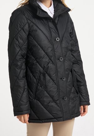 Frau trägt eine schwarze gesteppte Jacke mit Knöpfen und aufgesetzten Taschen vorne über einem weißen Hemd und beige-farbenen Hosen, steht vor einem weißen Hintergrund.