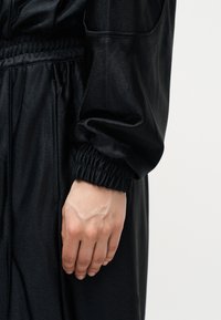 Laisva ranka šone, vilkinti juodą ilgomis rankovėmis drabužį su elastine rankogaliu ir tekstūruota medžiaga, fone – vientisa spalva.