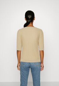 Mujer con el cabello oscuro en una coleta, vestida con un suéter de punto beige de mangas hasta el codo y jeans de mezclilla azules, de pie contra un fondo liso.