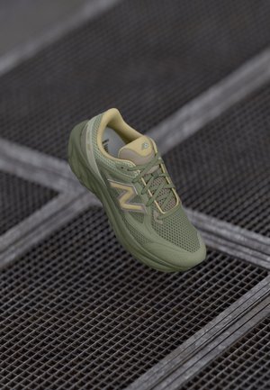 Jedna zielona tenisówka New Balance z ażurowym wzorem i sznurowadłami, spoczywająca na metalowej powierzchni z kratą.