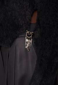 Antik guld brosch med två fåglar och hängande kristaller fästad på svart fluffig tyg över mörk texturerad klädsel.