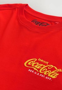 Czerwona bawełniana koszulka z żółtym haftowanym logo i napisem "Coca-Cola". Okrągły dekolt i krótkie rękawy, gładka faktura.