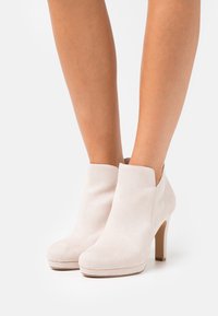 Un par de botas de tacón alto de ante color beige claro, usadas sobre piernas desnudas, contra un fondo blanco liso.
