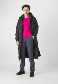 Cappotto lungo puffoso nero con cappuccio, indossato su una maglietta rosa brillante, abbinato a pantaloni blu scuro e scarpe nere. Presenta tasche con zip e un design elegante.