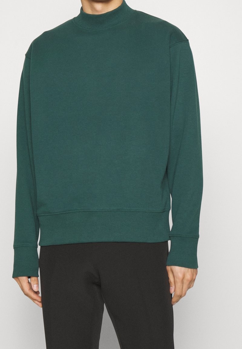 Homme portant un sweat à col montant vert forêt et un pantalon noir, debout devant un fond clair uni.