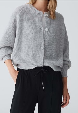 Femme portant un cardigan amples gris clair boutonné sur un haut blanc avec un pantalon noir à cordon de serrage, une main dans la poche, sur fond uni.