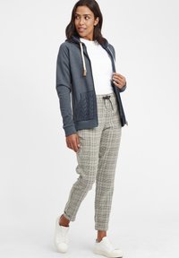 Hoodie bleu marine avec une poche frontale en tricot torsadé, cordons de couleur crème, t-shirt blanc et pantalon à carreaux gris. Des baskets blanches complètent la tenue.