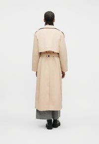 Άτομο με πλεγμένα μαλλιά που φοράει μπεζ παλτό τύπου trench, γκρι τζιν με φαρδιά πόδια και μαύρες χοντρές μπότες, στέκεται μπροστά σε απλό λευκό φόντο.