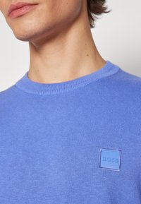 Gros plan d'une personne portant un pull en maille bleu avec un petit patch carré du logo indiquant "BOSS" sur la poitrine.