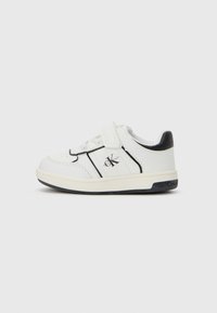 Baskets blanches pour tout-petits avec des accents noirs, bande Velcro, et logo Calvin Klein sur le côté et la semelle, sur fond blanc uni.