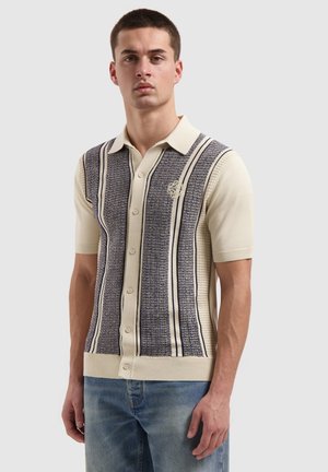 Junger Mann, der ein cremefarbenes kurzärmliges Hemd mit vertikalen, strukturierten grauen und schwarzen Streifen trägt, kombiniert mit blauen Jeans.