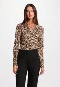 Femme portant une chemise en imprimé léopard avec un col, associée à un pantalon noir. Elle se tient devant un fond blanc uni.