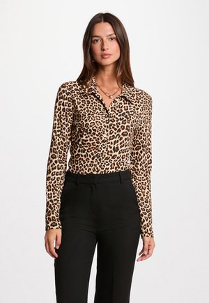 Femme portant une chemise en imprimé léopard avec un col, associée à un pantalon noir. Elle se tient devant un fond blanc uni.