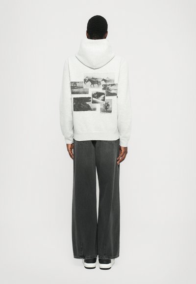 Haut à capuche gris clair présentant un collage d'images en noir et blanc au dos, associé à un pantalon large gris foncé et des baskets noires.