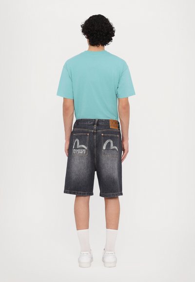 Camicia azzurra chiara a maniche corte, pantaloni corti di denim neri con cuciture a contrasto e dettagli del logo sulle tasche posteriori, abbinati a sneaker bianche.